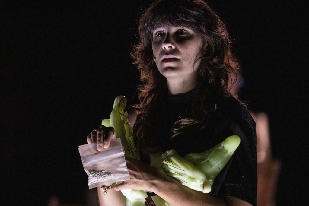 On voit Aurélia Lüscher un peu dans la penombre en train de monter un sachet en plastique contenant comme des petits objets metalique. Aurélia tiens aussi dans ses bras des statues flourescentes de la virge. Aurélia a les cheveux bouclés, longs aux epaules avec une frange. Elle porte unt-shirt.