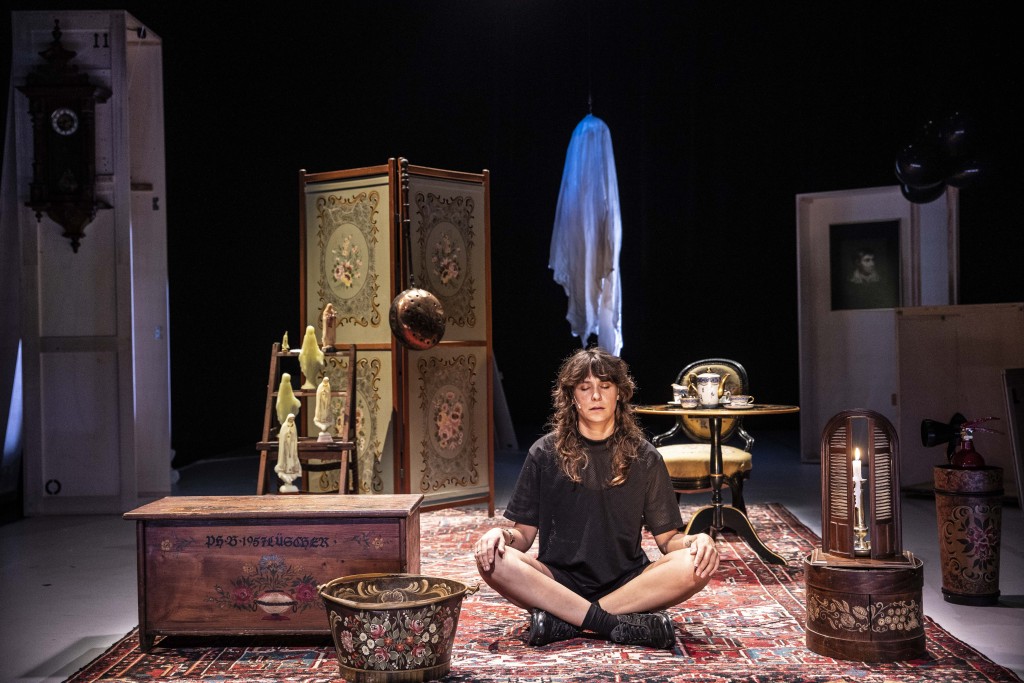 Aurélia Lüscher assise en tailleur sur un tapis. Elle a les yeux fermés comme si elle était en train de méditer. Autour d'elle des objets qui font un peu vieux salon.