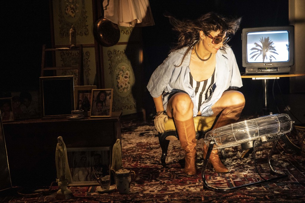 Aurélia Lüscher assise sur un petit tabouret avec des bottes qui lui arrivent presque au genoux. Elle regarde un ventilateur devant elle qui lui decoiffe les cheveux elle porte des lunettes de soleil.