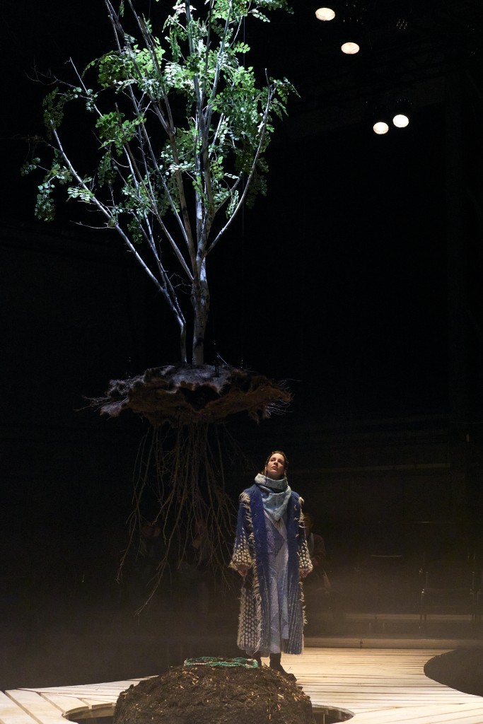 Un arbre suspendu par dessus une petite montagne de terre. Une personne l'observe. La tête en haut. Elle porte une longue robe et un long manteau.