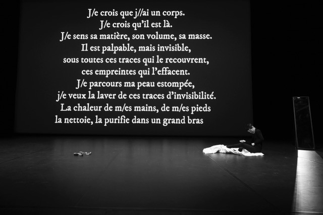 Un texte blanc sur un fond noir. Une personne assise à droiute. Le texte est propjeté sur un fond noir au fond du plateau.